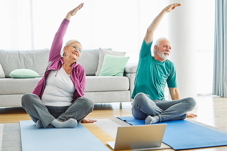 Dos personas adultas mayores realizando estiramiento sentados sobre el suelo. 