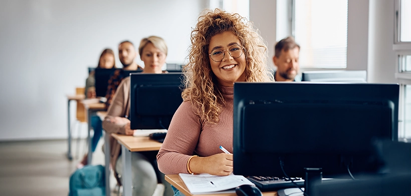 Mujer joven sonriente estudiando en la modalidad de bachillerato por ciclos frente a un computador, rodeada de compañeros de clase 