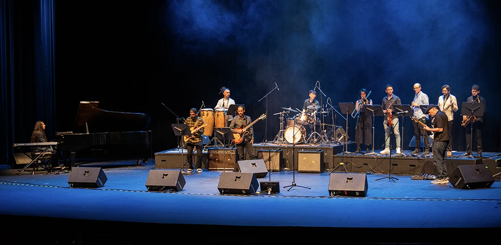 Agrupación musical en el escenario del Teatro Colsubsidio bajo luces azules, con teclado, guitarras, percusión e instrumentos de viento.